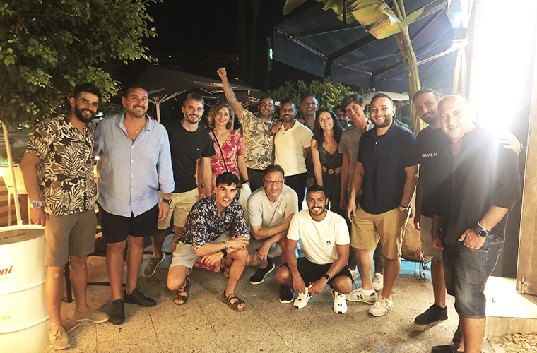 Un grupo de personas disfrutando de la cena de despedida en el campamento de pádel, un evento nocturno al aire libre, con atuendos veraniegos informales y un ambiente tropical iluminado.