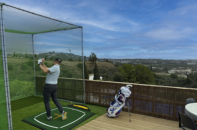 A person practicing golf inside the practice are with a net infront and a golf bag, the practice is taking place in wonderful view to the valley